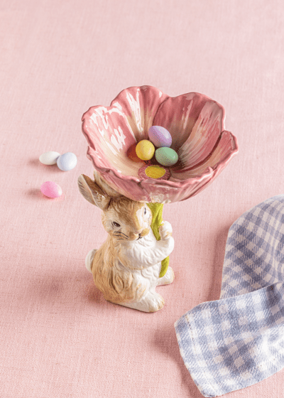 Decorative scene with a rabbit holding a pink flower-shaped bowl filled with Easter eggs on a pink background.