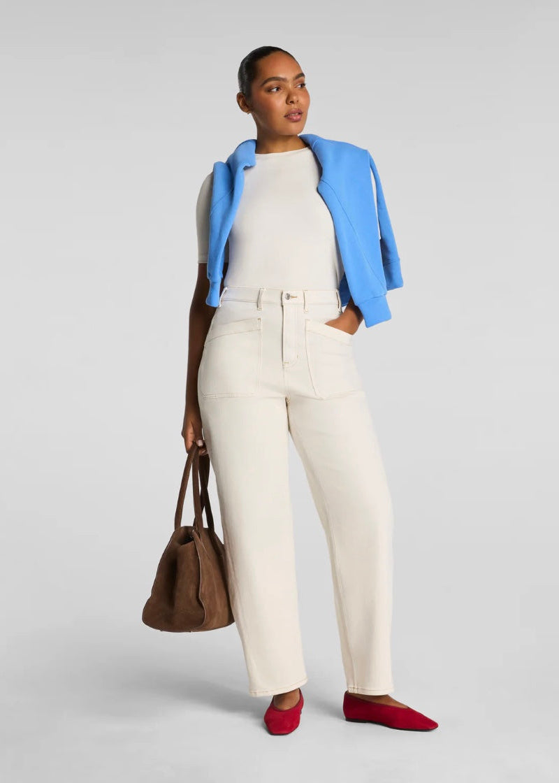 Woman wearing a blue jacket, white top, and cream pants holding a brown bag on a light gray background