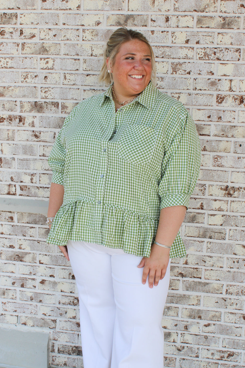 Woman wearing a green checkered shirt and white pants standing against a brick wall.