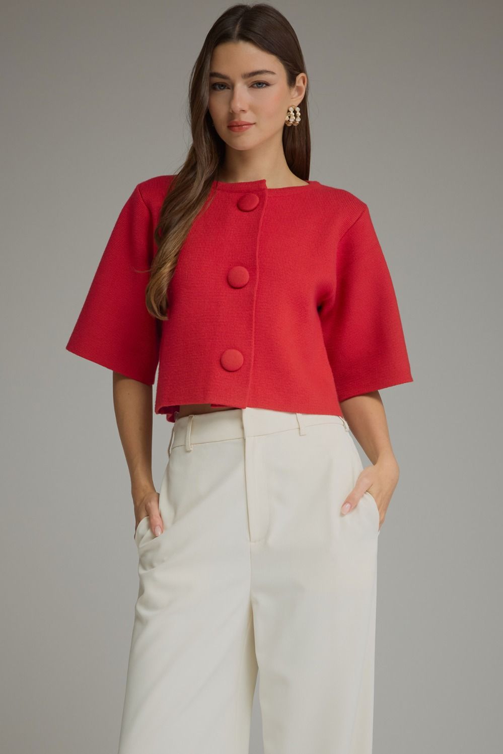 Red cropped short sleeve sweater jacket with oversized fabric-covered buttons, round neckline and structured knit, styled with ivory trousers.