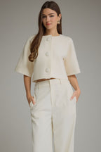 Ivory cropped sweater jacket with short sleeves, round neckline and statement covered buttons, styled with tailored trousers for a classic preppy look.